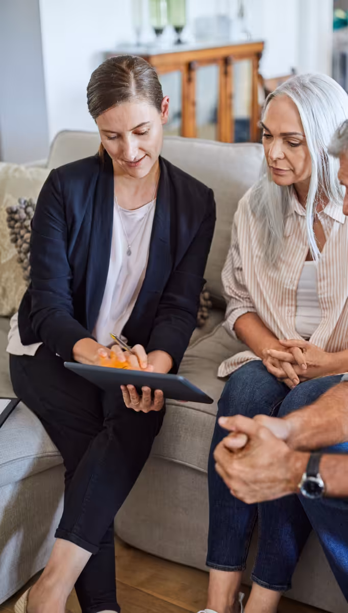 Two women sit on a sofa. The woman on the right is holding an iPad tablet that she and the second woman, who has grey hair and looks older, are both looking at.