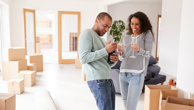 First-time home buyers celebrating their move into a new home, surrounded by moving boxes and raising glasses together.
